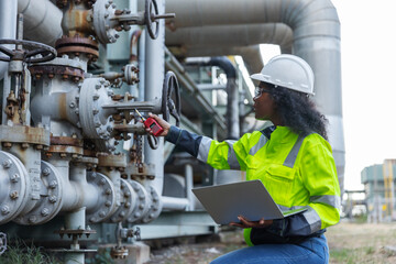 A female engineer wearing a safety helmet and high-visibility jacket works on a laptop beside an industrial pipeline system,highlighting engineering,technology, and maintenance in a complex industrial