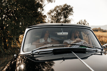black car, front view. There are two people sitting inside the car. A man is driving, he is looking towards the passenger seat. There's a woman lying in the back seat