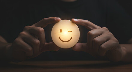 Hands holding a glowing wooden smiley face with a winking expression on a dark background.