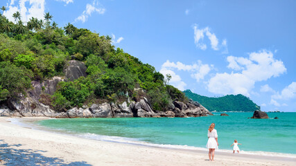 Koh Phangan, Thailand. A young woman in a white dress walks with her daughter on the coast of a beautiful beach.