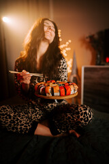 Woman enjoys sushi in bed wearing pajamas while surrounded by warm light at home