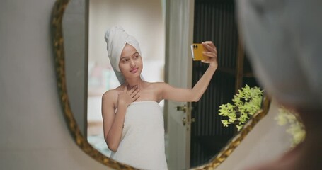Indian happy gen z young adult female lady wrapped towel head body after shower use mobile record video teen girl hold phone look cam mirror take selfie make pose enjoy fun joy day spend time house