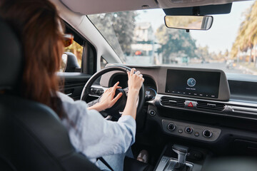 Woman driving a car with hands on the steering wheel, focused on the dashboard screen displaying...