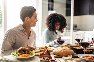 Close-up of friends laughing and enjoying a homemade meal together. Bright dining area with diverse young adults sharing food, wine, and conversation. Warm atmosphere promoting inclusion friendship