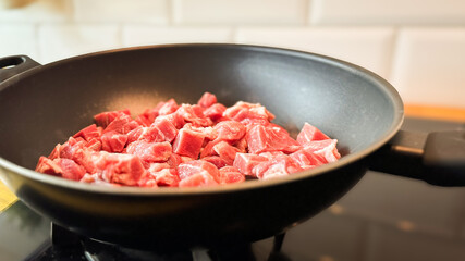 Fresh raw diced meat in a black non-stick frying pan on a modern kitchen stove, ready for cooking. Minimalist kitchen background with natural lighting