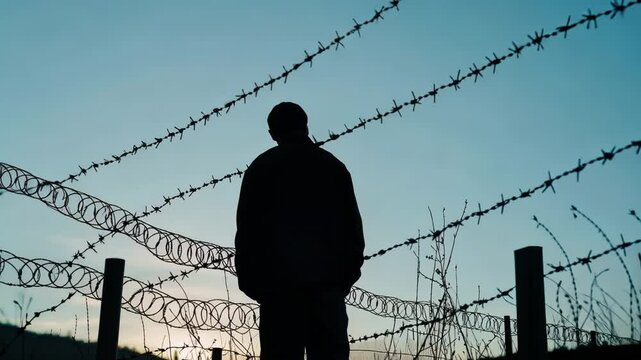 Man stands in front of a barbed wire fence. The sky is blue and the man is wearing a black jacket