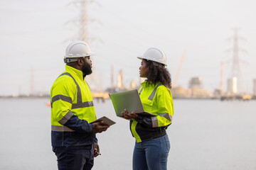 Two engineers in safety helmets and high-visibility jackets discuss work while holding a tablet and clipboard at a power plant site,with transmission towers and industrial structures in the background