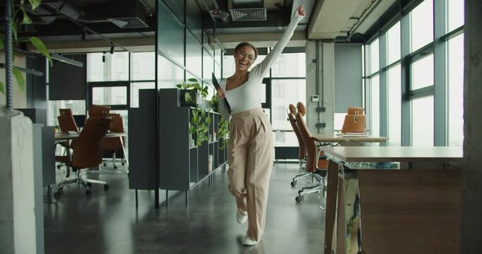 Happy young professional woman in stylish outfit celebrating success while holding a clipboard, standing in a modern office with plants, leather chairs, and large glass windows