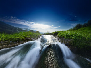 Serene mountain stream cascades over rocks, flowing into a lush green valley under a vibrant blue sky.  Sunlight filters through the clouds.