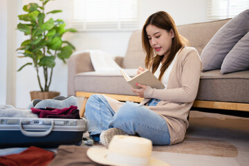 Asian woman planning her travel at home, ticking off a detailed packing list as she packs travel essentials into her suitcase.woman organizing everything she needs for a successful trip.