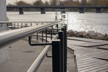 River bank with concrete promenade and metal railings. A bridge over the river can be seen in the background.