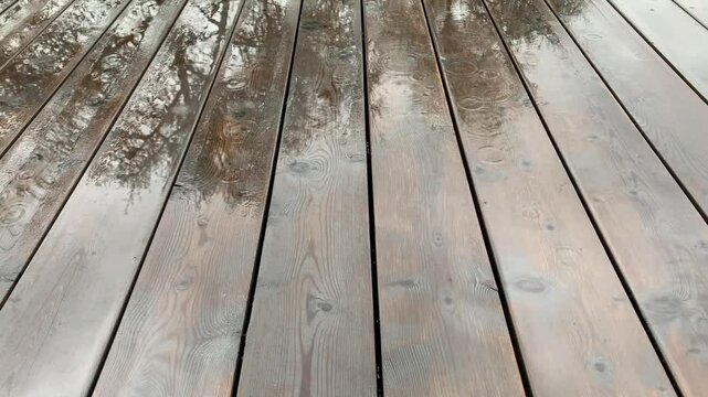 Wooden planks are dark brown and wet, with water droplets visible. The reflection of the trees and the sky can be seen in the wet surface of the deck