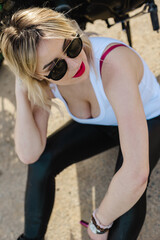 A stylish woman with blonde hair sits on the ground wearing sunglasses, red lipstick, a white tank top, and black pants. The photo captures a confident, relaxed, and fashionable vibe