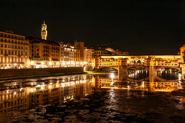 Fototapeta premium Night falls over Florence, illuminating the Ponte Vecchio and surrounding buildings. The tranquil Arno River reflects the vibrant lights, creating a magical atmosphere in Tuscany.