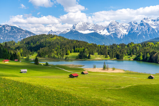 Geroldsee lake in Bavaria, Germany, featuring rustic wooden huts and the majestic, snow-dusted Karwendel mountains under a sunny blue summer sky