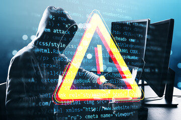 Hacker in hoodie working on a computer with digital warning sign overlay and code in background.
