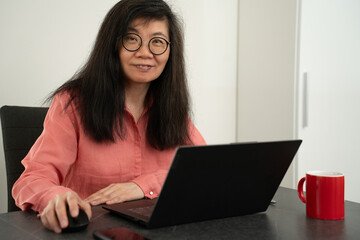 Naklejka premium Middle aged chinese business woman with glasses, long black hair and red blouse is working with a laptop inside an office, just looking up from her work. Red cup of coffee near by.