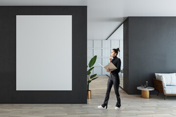 Modern office interior with a businesswoman holding a folder near a large blank poster on the dark wall, wooden floor, and clean design.