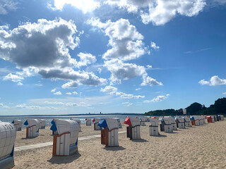 An idyllic Baltic Sea beach under a wide, blue sky with white clouds. Beach chairs in red, blue and white are lined up along the fine sand, the coast stretches out on the horizon. Pelzerhaken, Neustad