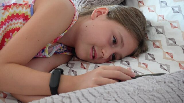 Portrait of a smiling preteen girl lying on the bedspread in a bed using her smartphone
