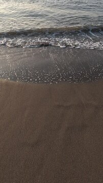 close-up view of ocean waves gently rolling onto a sandy beach. The foamy white water contrasts with the brown wet sand as the tide ebbs and flows along the shoreline. Slow motion vertical video
