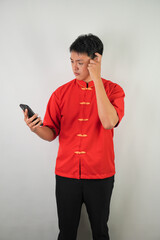 Confused young asian man wearing red traditional chinese clothing is calling and arguing to someone using his smartphone, isolated over white background. Concept for Lunar New Year Party.