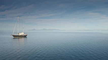 Fototapeta premium Wooden yacht anchor peacefully on serene tropical sea, with distant island visible on horizon under cloudy blue sky. Remote wild nature paradise, exotic summer travel destination. Drone flight