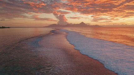 Ocean waves gently rolling sand shore of Tahiti tropical island at sunset. Vibrant orange colors and dramatic clouds reflecting in water. Remote wild nature paradise, exotic summer travel. Aerial shot