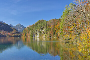 The serene waters of the Alpsee perfectly reflect the vibrant autumn foliage of the surrounding Bavarian Alps. A breathtaking glimpse of the combination of majestic mountains, forests and water.