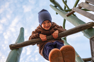 Cheerful child in warm clothing and striped beanie on wooden playground structure with sky in...