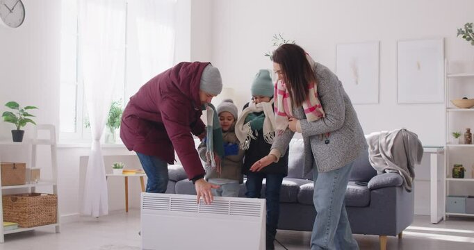 Freezing young family wearing warm winter jackets standing near electric heater. Parents and children suffering from low temperature, trying to warm up during cold winter and facing heating problems.
