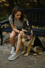 A happy young woman strokes her beloved dog while sitting on a wooden bench in a summer park. An intelligent and obedient German Shepherd spends time outside with a teenage owner.