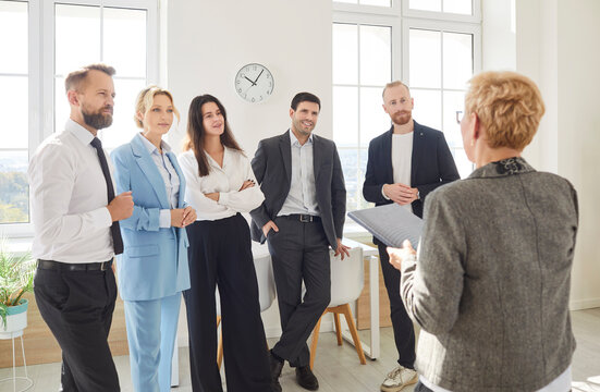 Back view of business leader or coach standing in front of diverse group of company employees. Team of colleagues standing in office, listening to female colleague during business meeting or training.