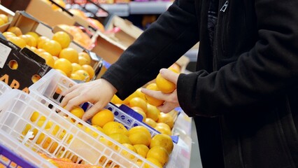 Mature shopper hand picking ripe tangerines from colorful produce display, selecting vitamin rich...