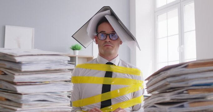 Confused tired employee bounded with yellow tape sitting at work desk and looking unhappily at stack of paperwork. Frustrated office worker with folder with papers on his head stuck on work forever.