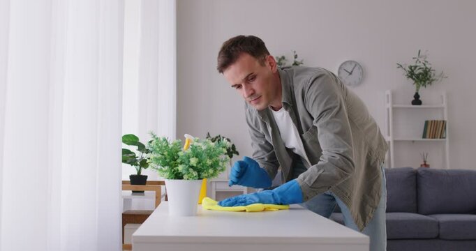 Happy cheerful male young cleaner wiping desk with household detergent supplies at home. Smiling male person from cleaning service washing job workplace and cleaning house. 4k video. - Powered by Adobe