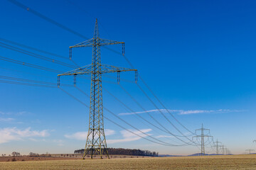 Ein Hochspannungsmast in einer frostigen Winterlandschaft, umgeben von Stromleitungen, Feldern und einem klaren blauen Himmel. Die Szene thematisiert die Energie&uuml;bertragung in der kalten Jahreszeit.