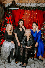 Naklejka premium Four women in stylish outfits posing in a festive Christmas setting with decorated trees, lights, and wrapped gifts in the background.