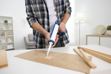 Man with caulking gun glueing plywood at white table indoors, closeup