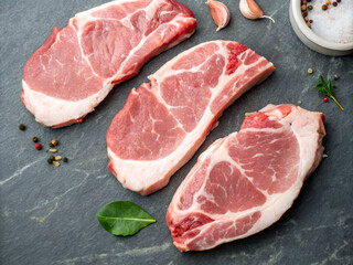 Fototapeta premium slices of uncooked pork chop on a dark slate cutting board, garnished with a few rosemary sprigs. The background is a rustic, weathered gray surface, and there are other rosemary sprigs and a spice c