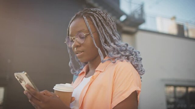 Camera moving around woman with braided hair dyed light purple. Female smiling while looking at smartphone display. Typing with one finger on touch screen. Wearing round glasses with golden frames.
