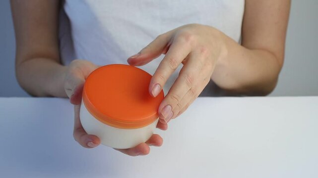 Caucasian woman european opens a jar of hand cream and applies the cream. White background. Close-up. Copy space. White packaging with orange cap