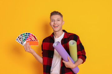 Young decorator with color palettes and rolls of wallpaper on orange background