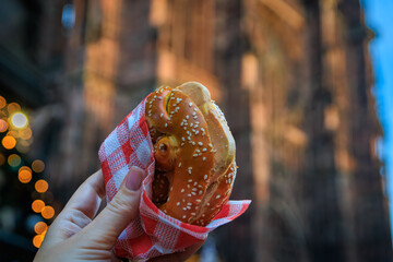 Woman holds a foie gras pretzel sandwiches at the Chrismas Market in Strasbourg, France with the Notre Dame Cathedral in the background