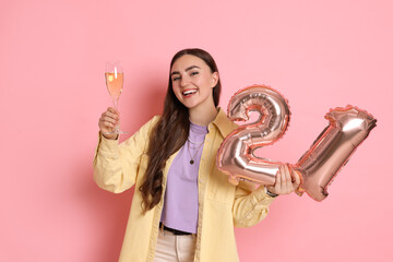Coming of age party - 21st birthday. Happy young woman with number shaped balloons and glass of sparkling wine on pink background