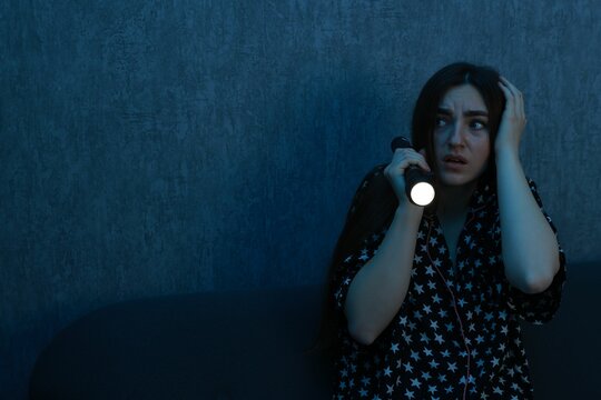 Fear of darkness. Scared young woman with flashlight on couch indoors at night, space for text