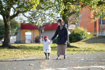 Fototapeta premium A 3-year-old Japanese girl wearing a red knitted hat and a white down jacket walks with a woman in her 30s through fallen leaves in a park in Hakata, Fukuoka Prefecture, on a cold winter afternoon.