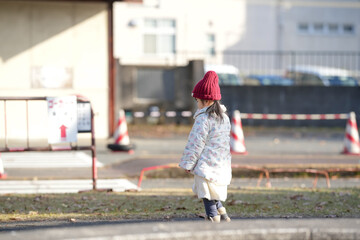 A 3-year-old Japanese girl wearing a red knitted hat and white down jacket walks through a park in...