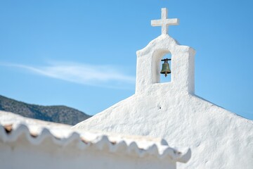 Fototapeta premium A charming white bell tower rises against the backdrop of a clear blue sky. The structure features a classic cross and a rustic bell, showcasing traditional architecture.