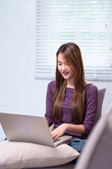 A young woman working on a laptop while sitting on a sofa, smiling and appearing focused, in a cozy home setting